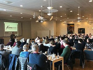 Personer sitter runt bord och lyssnar på en föreläsning.