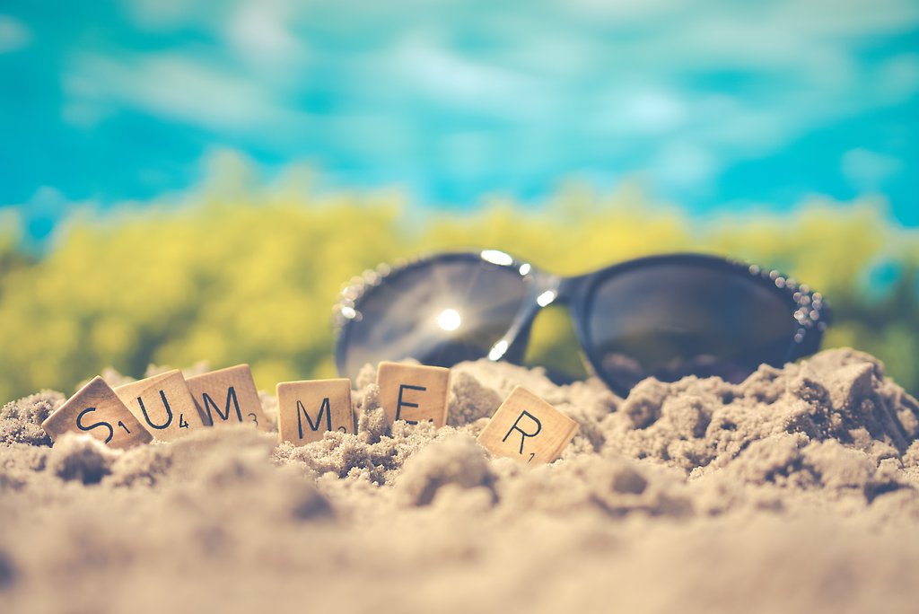Närbild på ett par svarta solglasögon som ligger på en strand med en text "summer" i träklossar.