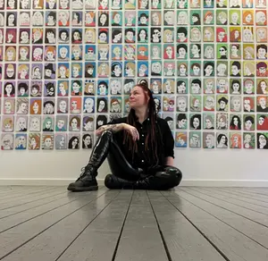 Kvinna sitter nedanför massa små färgglada porträtt som hänger bredvid varandra.
