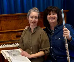 Två kvinnor sitter bredvid varandra. Den ena håller i en tvärflöjt och den andra i en bok.