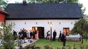 Ett vitt stenhus med svart tak. Framför huset minglar festkädda människor.