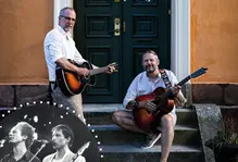 Två män framför en orange husfasad med varsin akustisk gitarr.