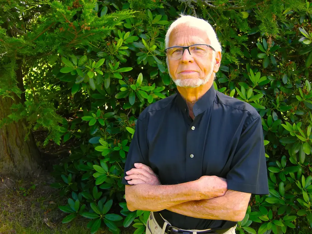 Äldre man med vitt hår och vitt skägg och glasögon som står framför ett grönt buskage och tittar in i kameran.
