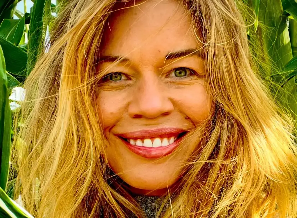 Leende kvinna med ljust hår står framför gröna växter.