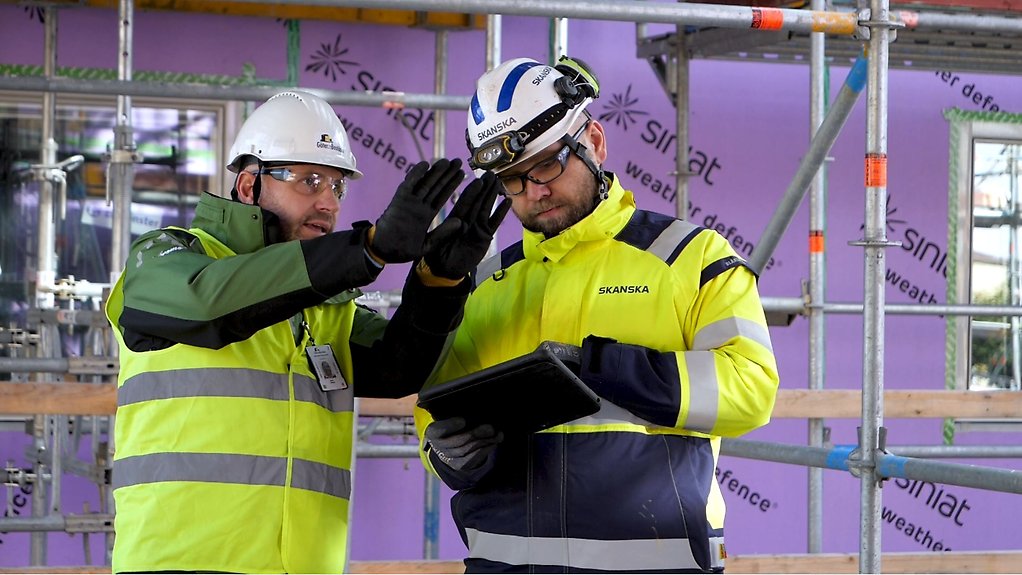 Två män i reflexvästar och bygghjälmar framför ett bygge. En håller visionärt upp händerna.