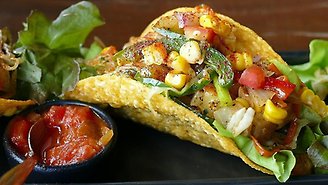 Svart bakgrund med ett gult tacobröd fylld med olika grönsaker som gul majs, röd tomat, grön gurka och sås över.