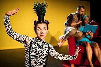 En kvinna i zebratröja balanserar en krukväxt på huvudet. I bakgrunden sitter en man och en kvinna i en fåtölj och läser en bok.