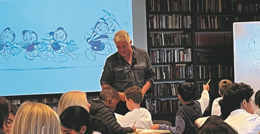 Fotot visar en man med grått hår som står framför en skolklass som får lära sig att teckna serier. På en skärm bakom mannen syns Kalle Ankafigurer i svartvitt.