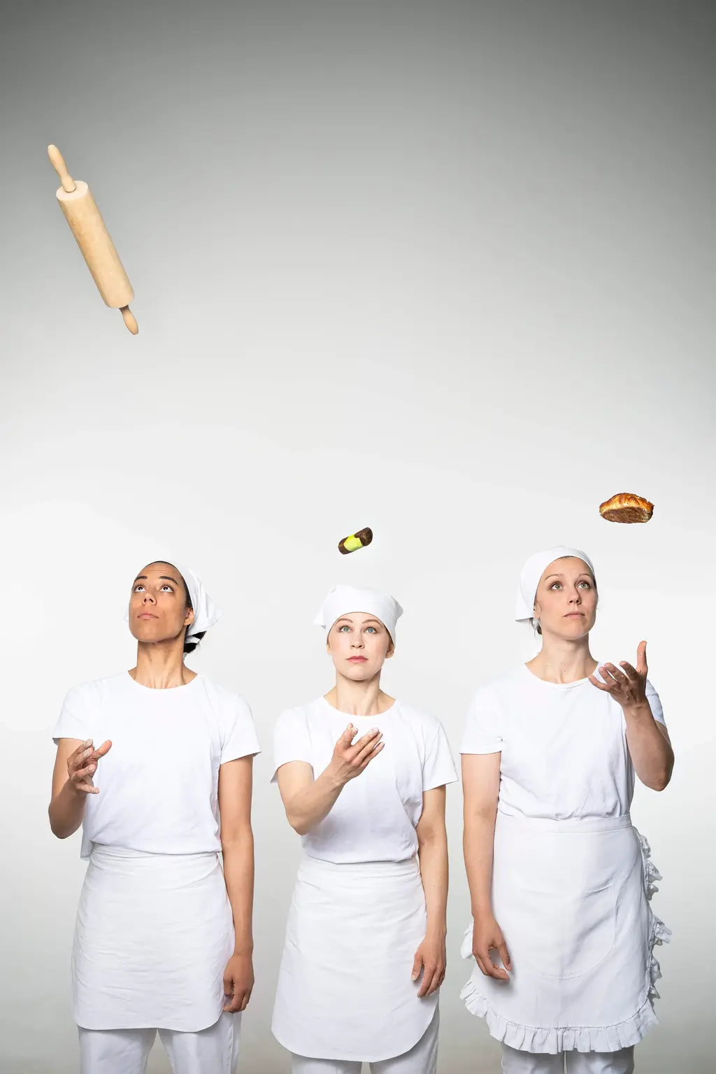 Tre kvinnor står på rad i bagarkläder och bagarmössor och jonglerar med kakor och en brödkavel.