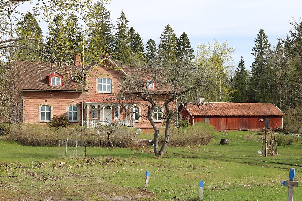 En bild som visar ett hus i rött tegel och ett rödmålat uthus, en trädgård med träd och gräsmatta.