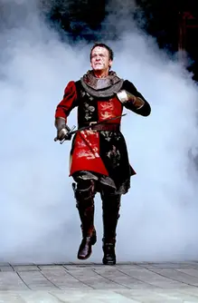 En blodig man med svärd och riddarutrustning springer ut ur dimman på en scen.