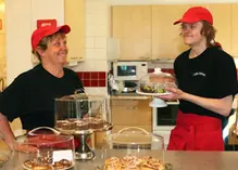 En kvinna och en man som arbetar i ett café. Framför dem syns bullar och kakor på olika fat.