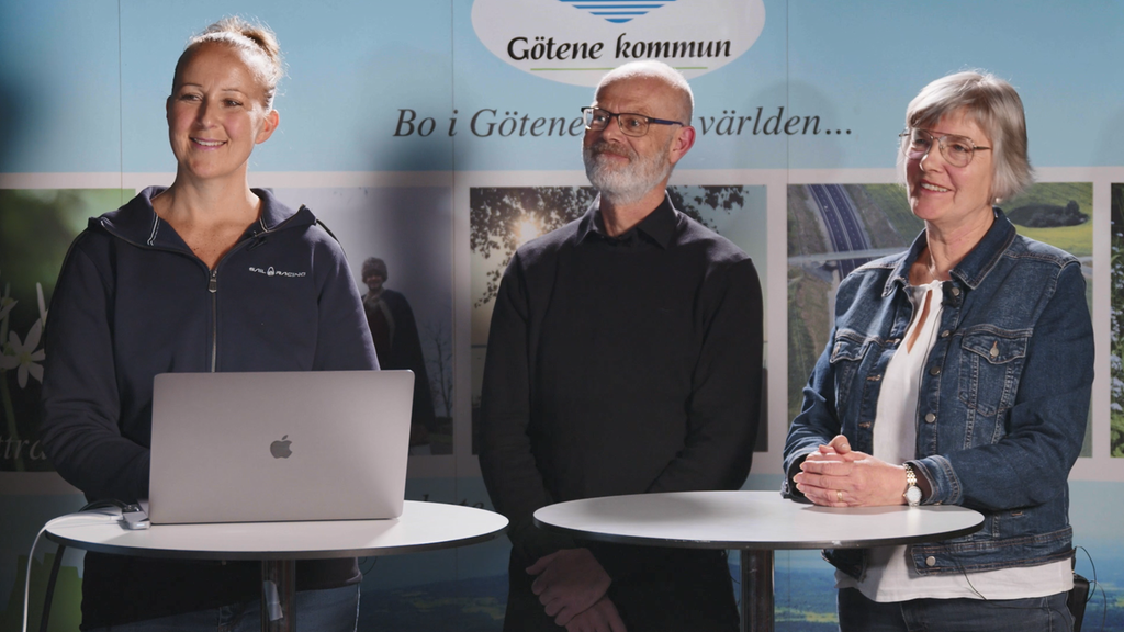 Tre personer står på rad bakom två, runda bord och håller i en presentation. 