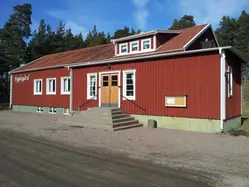 Bild på Holmestad Bygdegård. Avlångt rött hus med vita knutar. Trappa Burspråk på taket. Vit text på vänster framsida "Bygdegård".