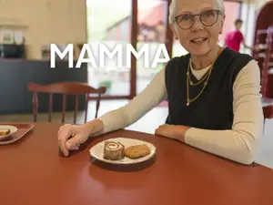 En kvinna som sitter vid ett bord med en kaffekopp. Bredvid syns texten "mamma"